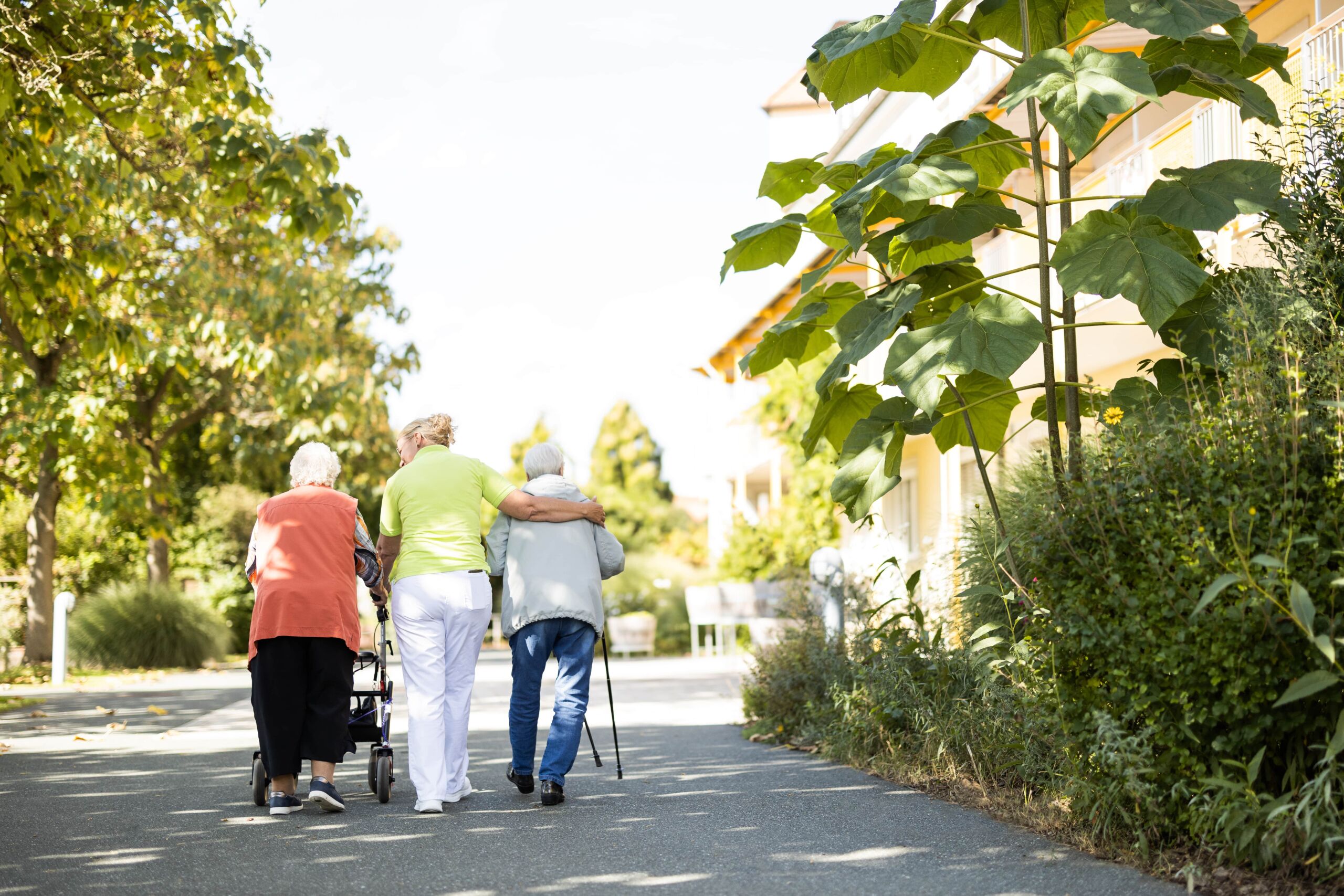 Pflegekraft des Seniorenhaus Menda begleitet zwei Seniorinnen bei einem Spaziergang durch den sonnigen Garten. Eine Seniorin nützt einen Rollator, die andere geht mit Gehstock und wird von der Pflegekraft stützend am Arm gehalten. Der Weg ist von großen Bäumen und üppigem Grün umgeben.