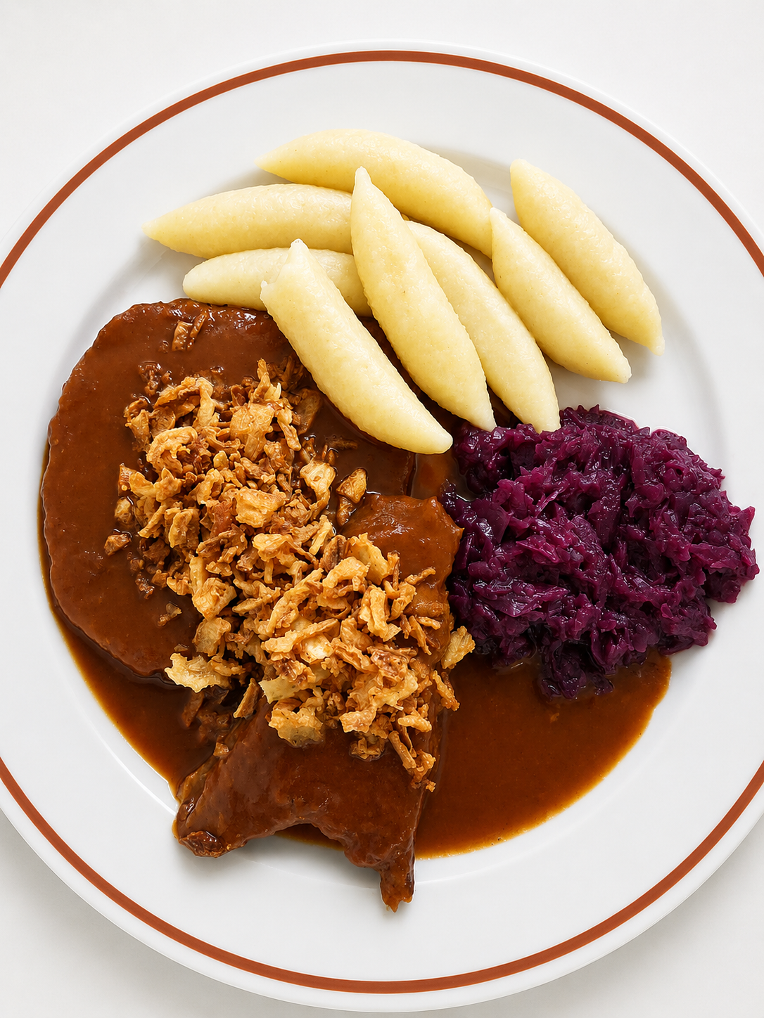 Rindsroulade mit dunkler Sauce, knusprigen Röstzwiebeln, weißen Nockerln und Rotkraut auf einem weißen Teller mit braunem Rand angerichtet.