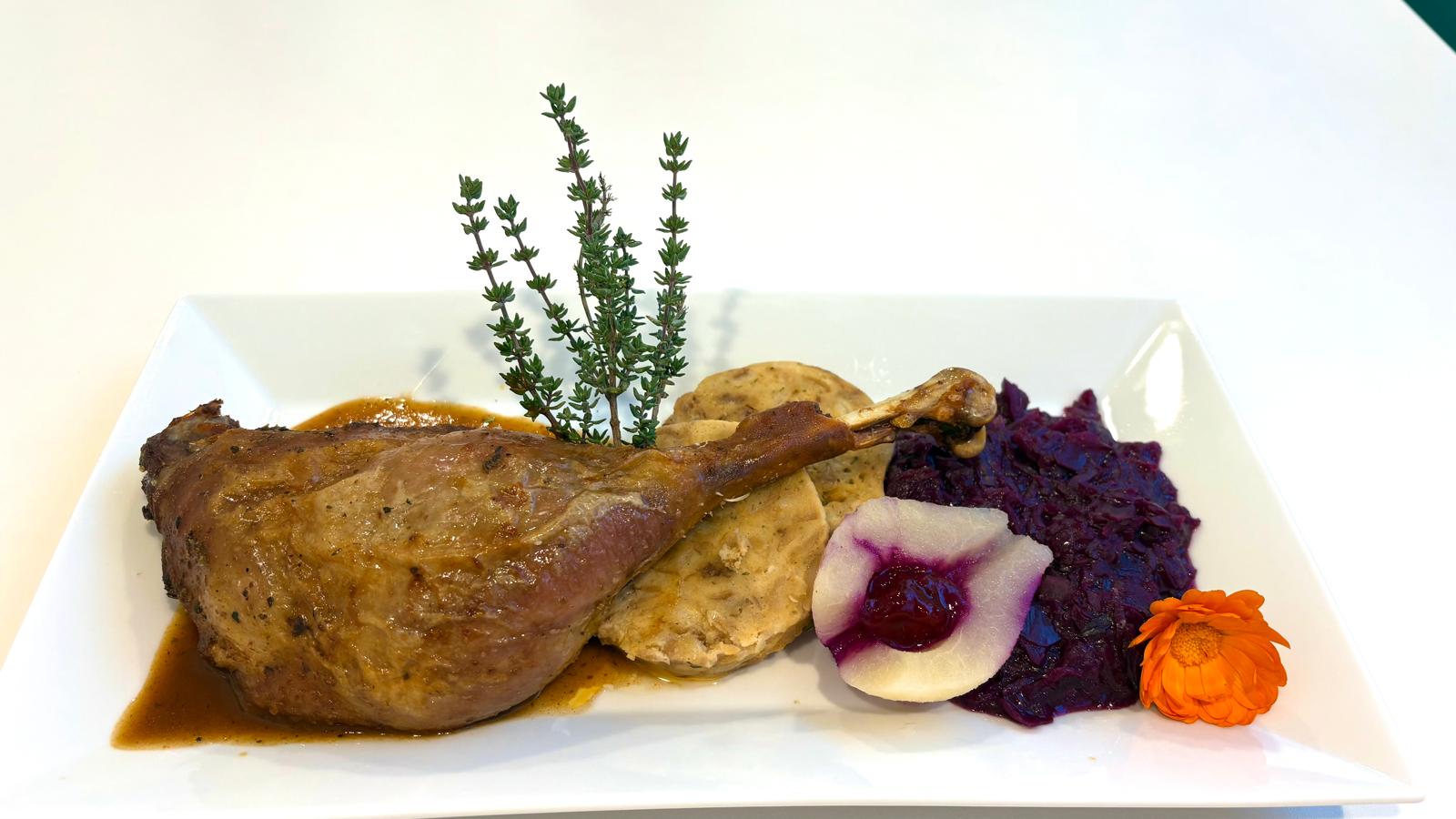 Gebratene Entenkeule mit Serviettenknödel, Rotkraut und dekorativer Blüte auf einem modernen weißen Teller angerichtet.