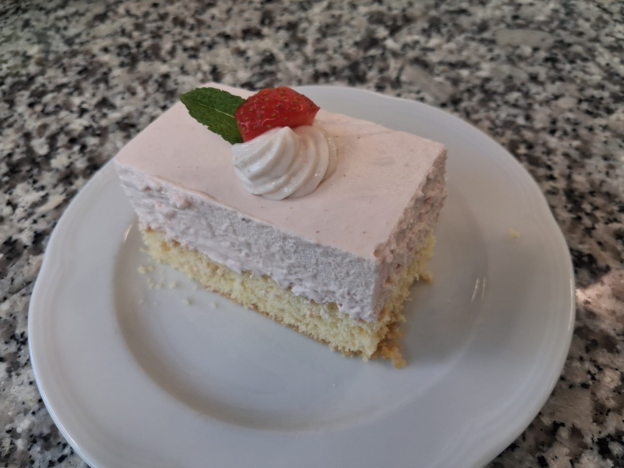 Luftige Erdbeer-Cremeschnitte mit hellem Biskuitboden, dekoriert mit Schlagobers, Erdbeere und Minzblatt auf weißem Teller.