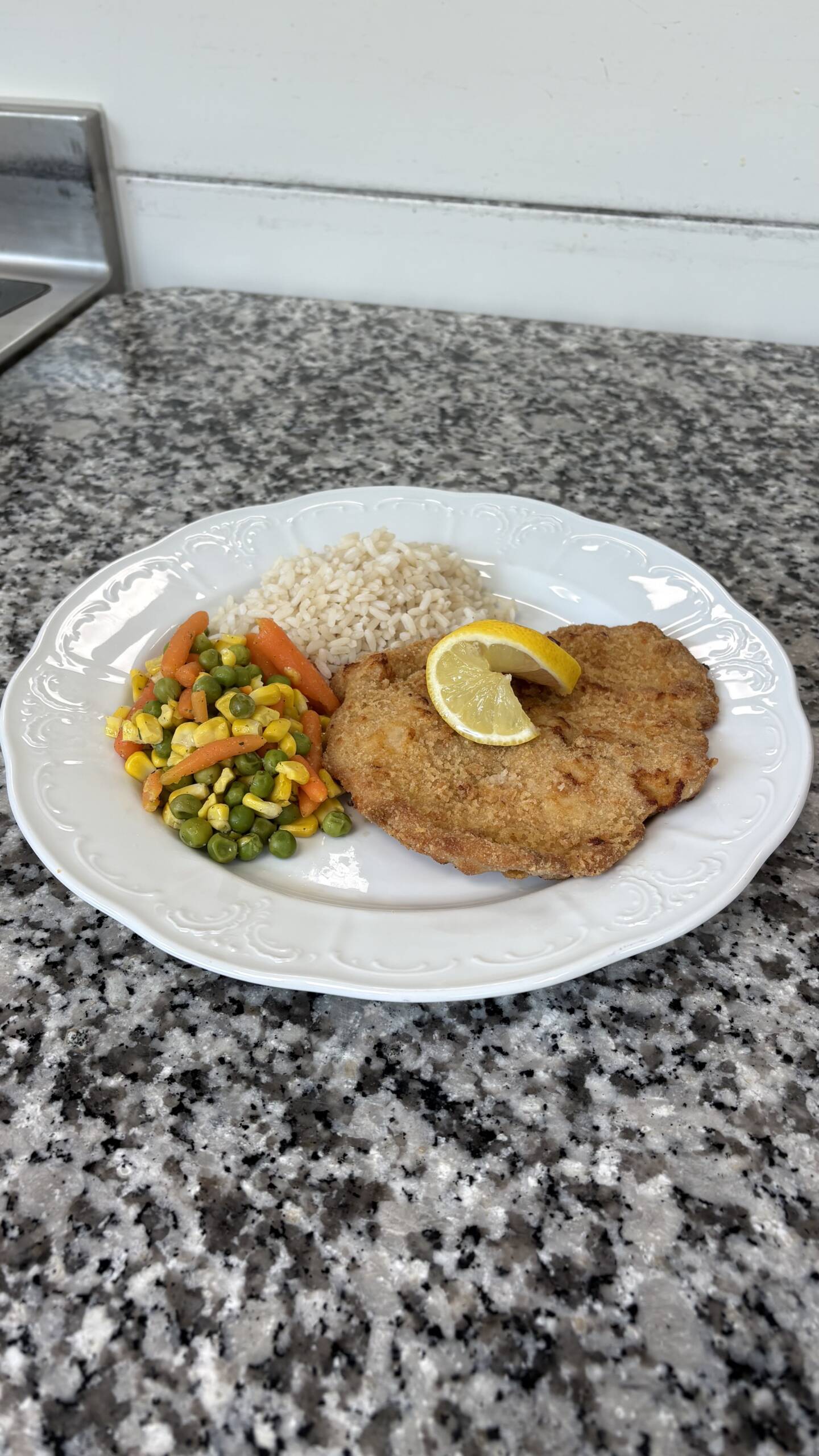 Goldbraun paniertes Schnitzel mit Zitronenscheibe, Reis und buntem Gemüse aus Erbsen, Karotten und Mais auf einem weißen Teller.