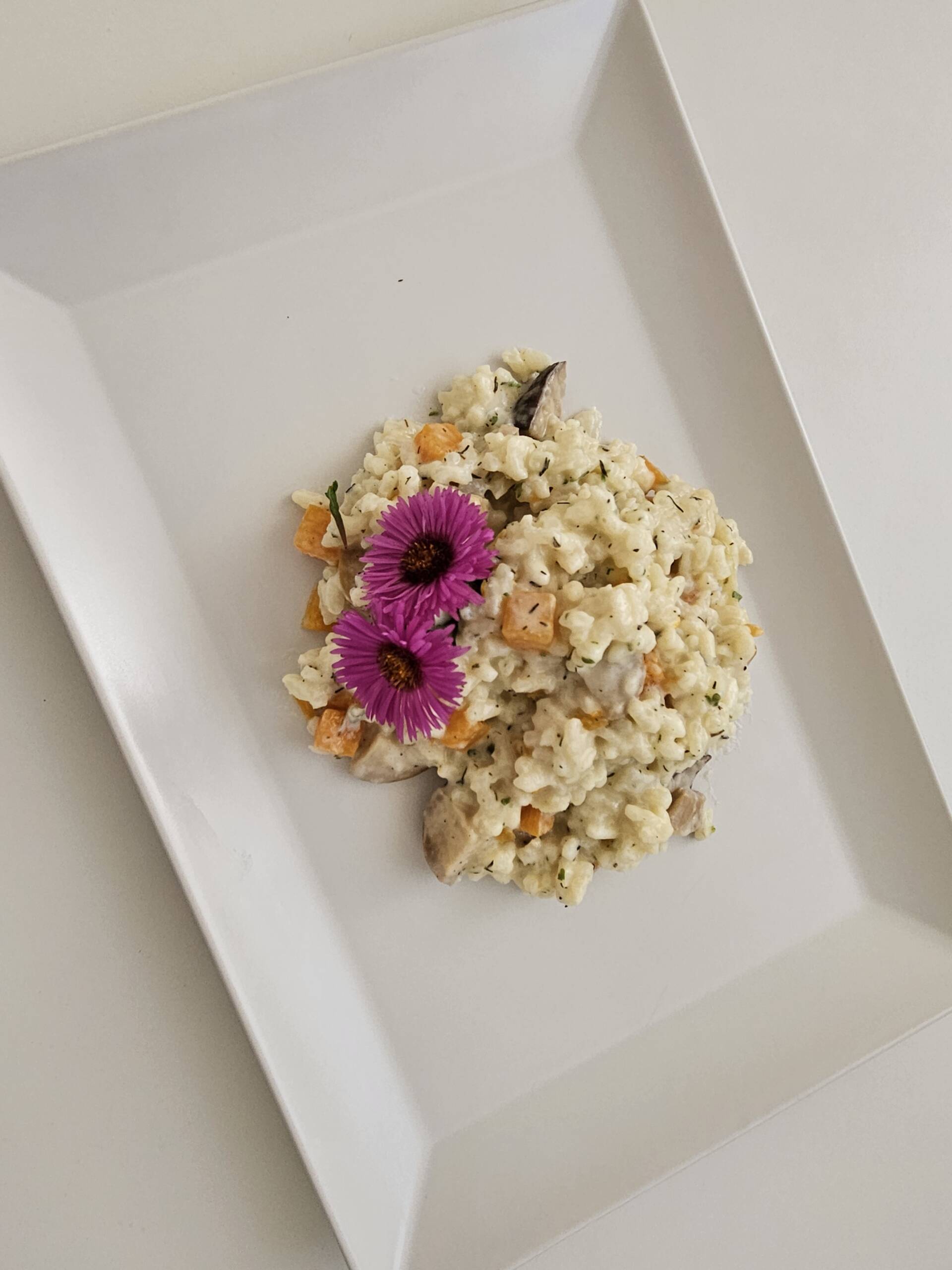 Draufsicht auf herbstliches Risotto mit Kürbis, Pilzen und essbaren Blüten auf eckigem Teller