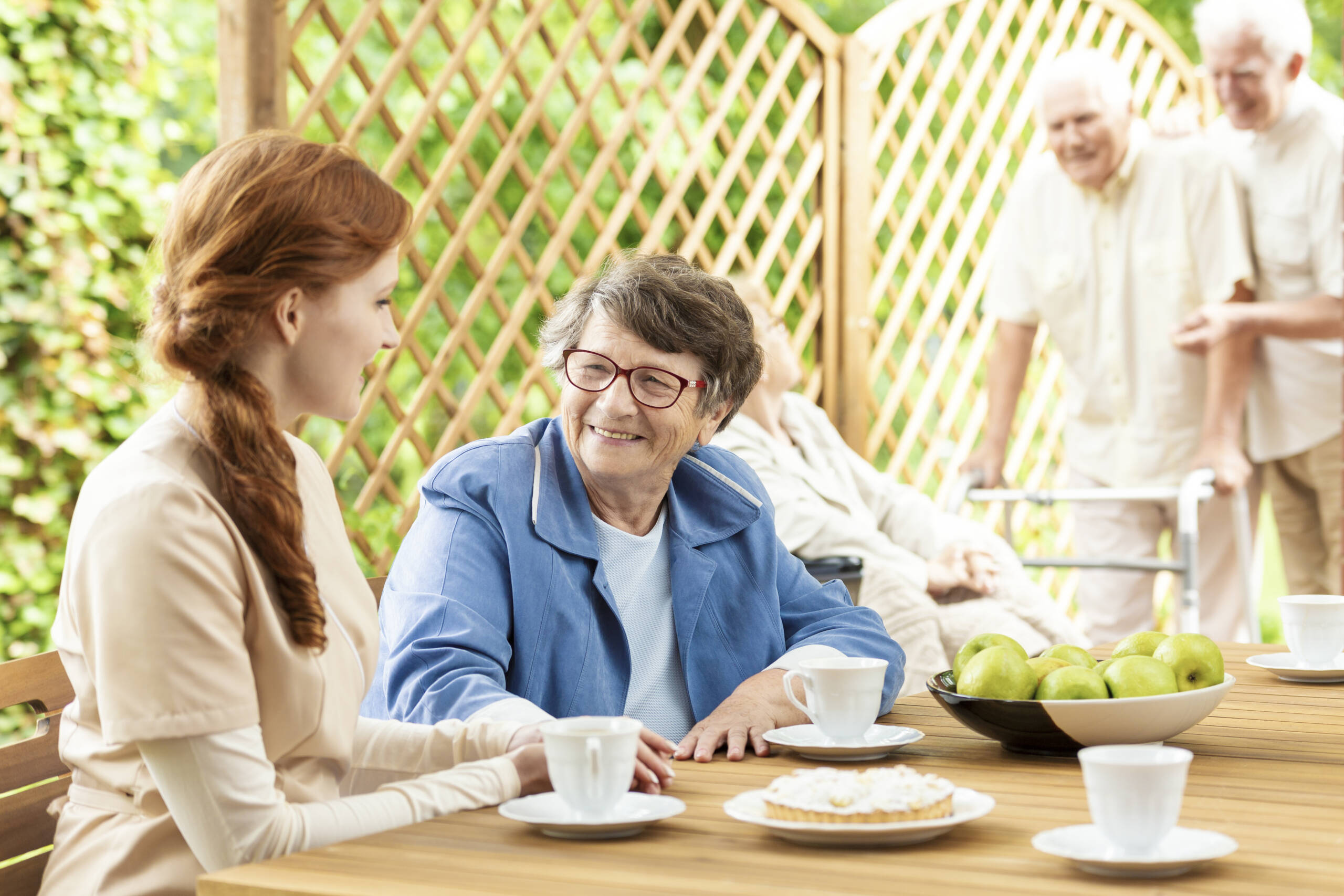 Seniorin und Besucherin sitzen lächelnd bei Kaffee und Kuchen auf der Terrasse im Grünen