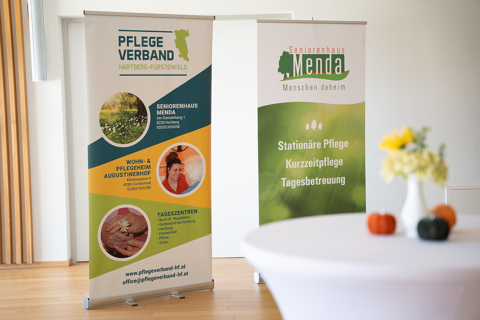 „Gold Standard“: Seniorenhaus Menda erhält das Weitblick-Zertifikat 9 Zwei Roll-up-Banner im Veranstaltungsraum: links vom Pflegeverband Hartberg-Fürstenfeld, rechts vom Seniorenhaus Menda mit „Stationäre Pflege, Kurzzeitpflege, Tagesbetreuung“. Im Vordergrund ein Stehtisch mit Blumen und kleinen Kürbissen.