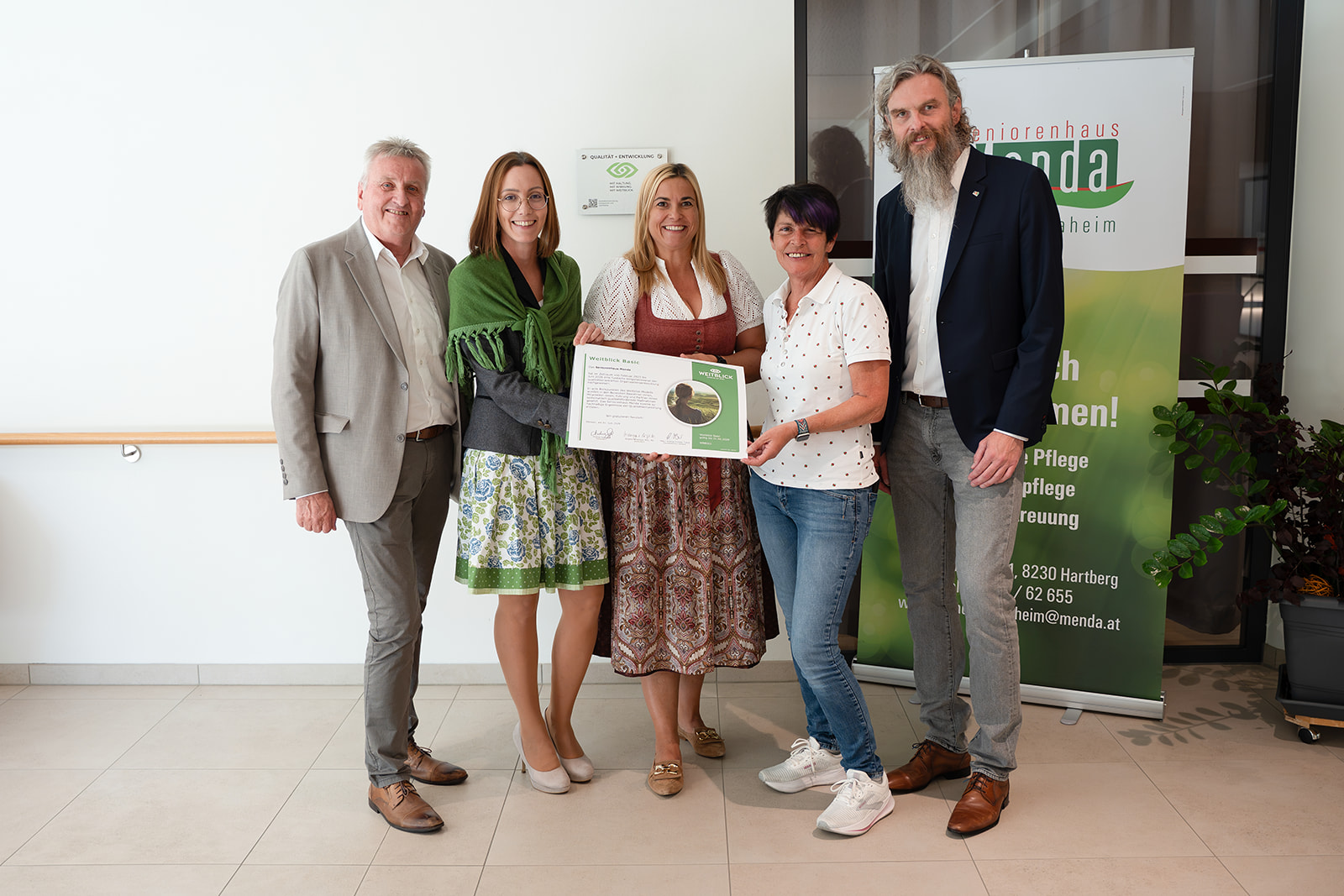 Gruppenfoto mit fünf Personen, die das Weitblick-Zertifikat halten, vor dem Menda-Roll-up und der neuen Plakette an der Wand. Gruppenfoto mit fünf Personen, die das Weitblick-Zertifikat halten, vor dem Menda-Roll-up und der neuen Plakette an der Wand.