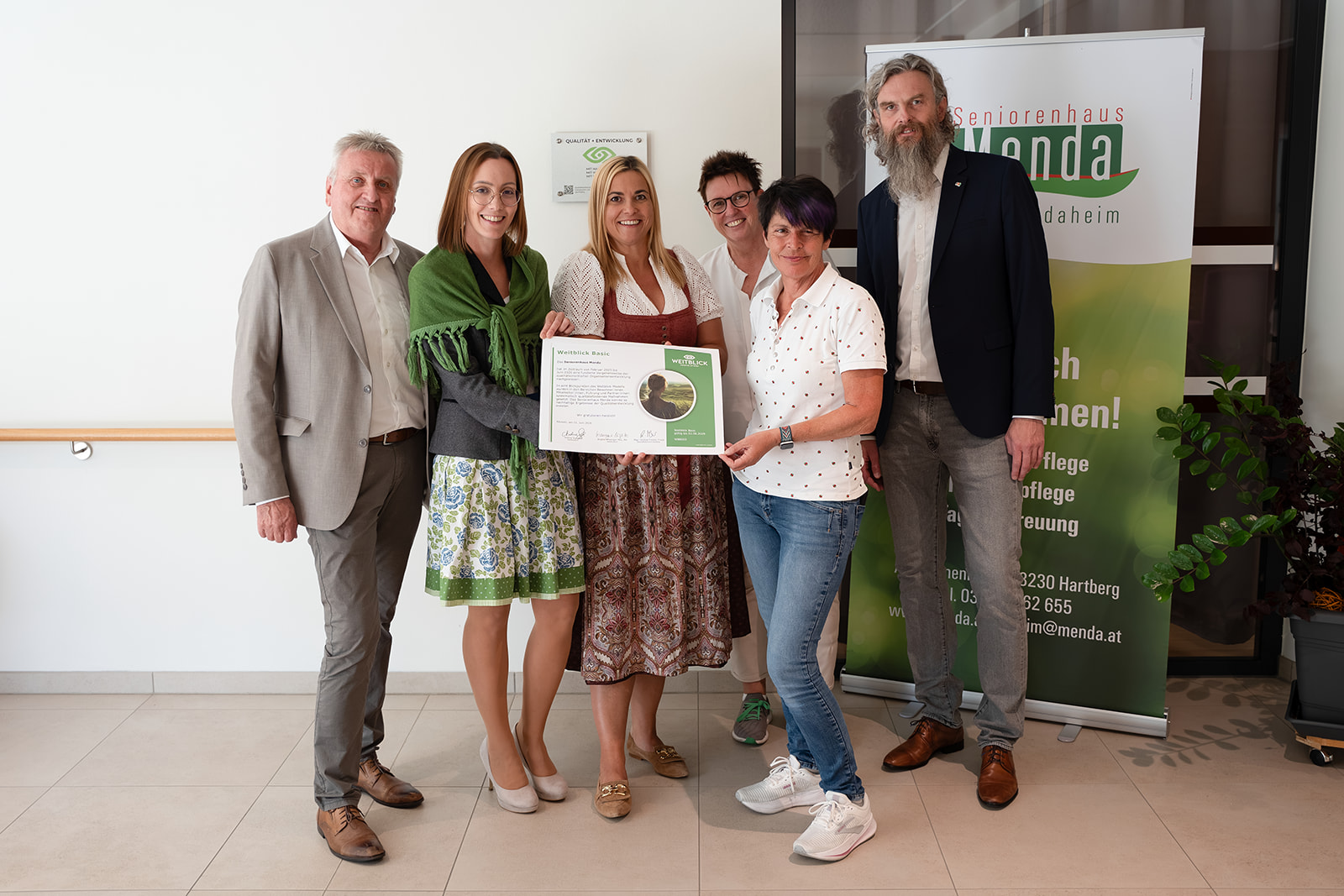 „Gold Standard“: Seniorenhaus Menda erhält das Weitblick-Zertifikat 4 Gruppenfoto mit sechs Personen, die das Weitblick-Zertifikat halten, vor dem Menda-Roll-up und der neuen Plakette an der Wand.