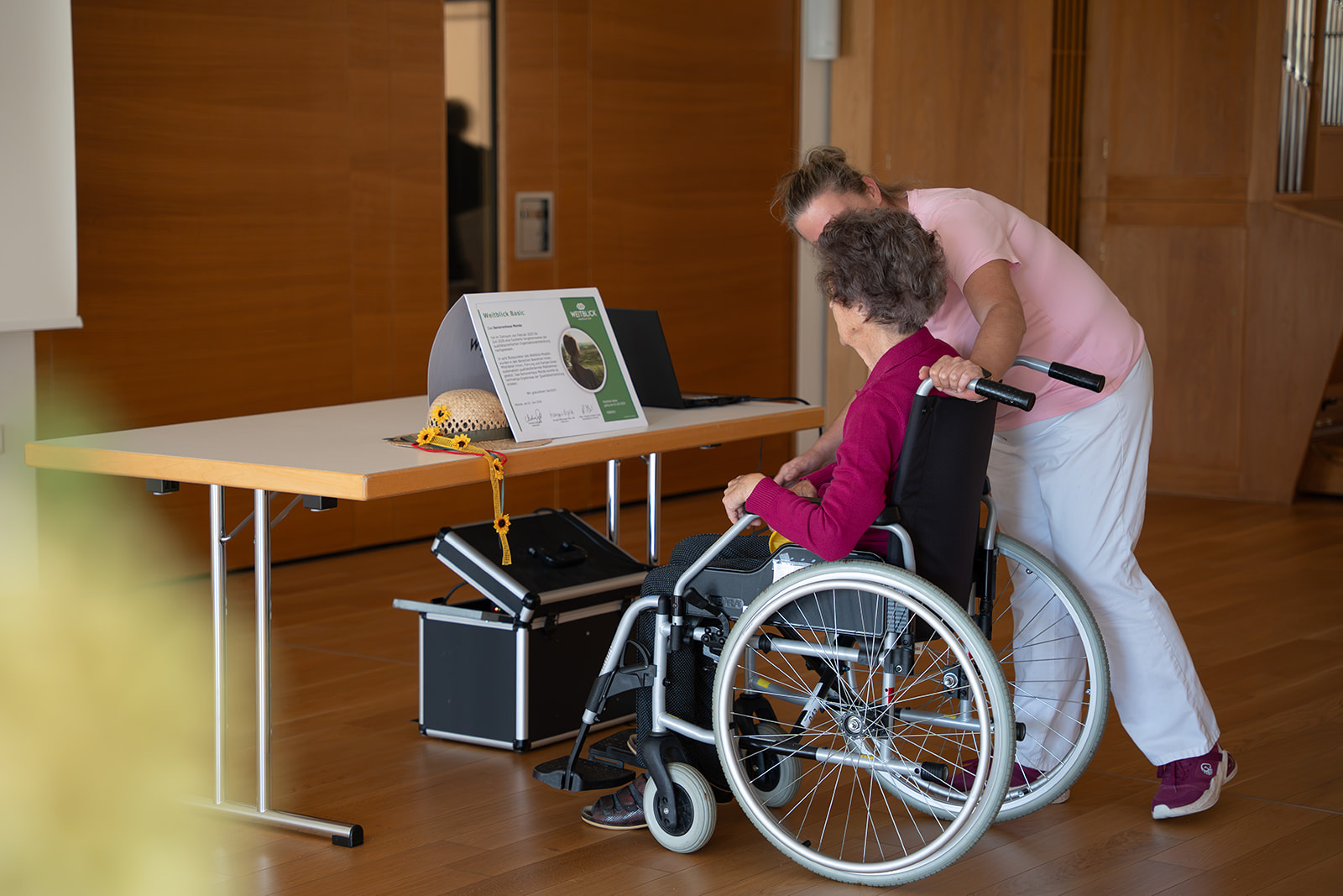 Eine Pflegerin beugt sich zu einer Bewohnerin im Rollstuhl. Gemeinsam betrachten sie das Weitblick-Zertifikat auf dem Tisch.