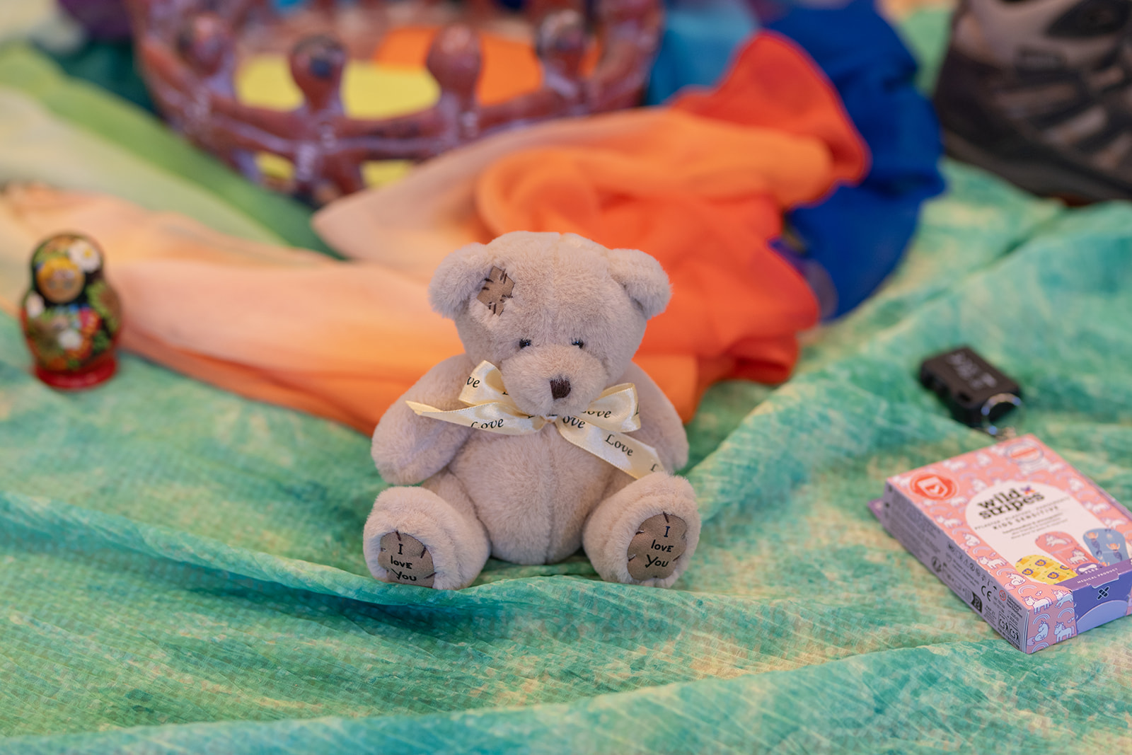 Kleiner beiger Teddybär mit aufgenähtem Flicken am Kopf, cremefarbener Schleife mit „Love"-Aufdruck und „I love you" auf den Fußsohlen sitzt auf einem grünen Tuch. Daneben liegen eine Packung bunte Kinderpflaster, eine Matroschka-Puppe und farbige Tücher.