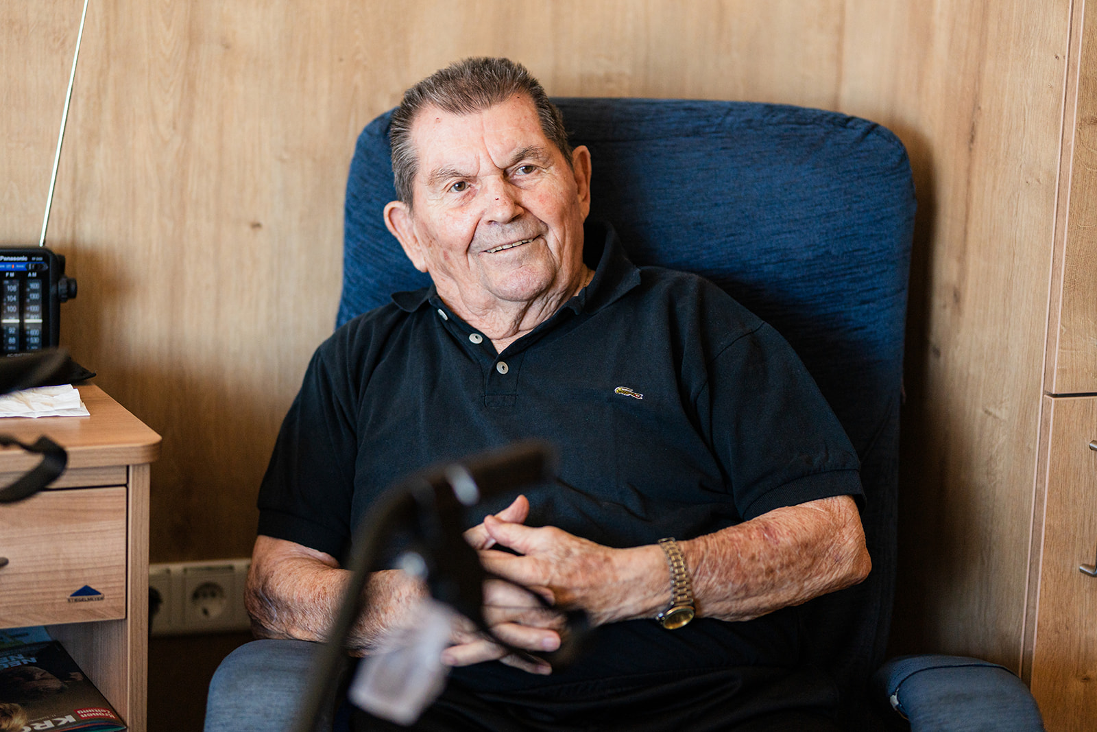Älterer Herr mit zurückgekämmtem grauem Haar sitzt lächelnd in einem blauen Polstersessel. Er trägt ein schwarzes Poloshirt und eine Armbanduhr, die Hände sind im Schoß gefaltet. Links steht ein Nachttisch mit Radio, im Vordergrund unscharf der Griff einer Gehhilfe.