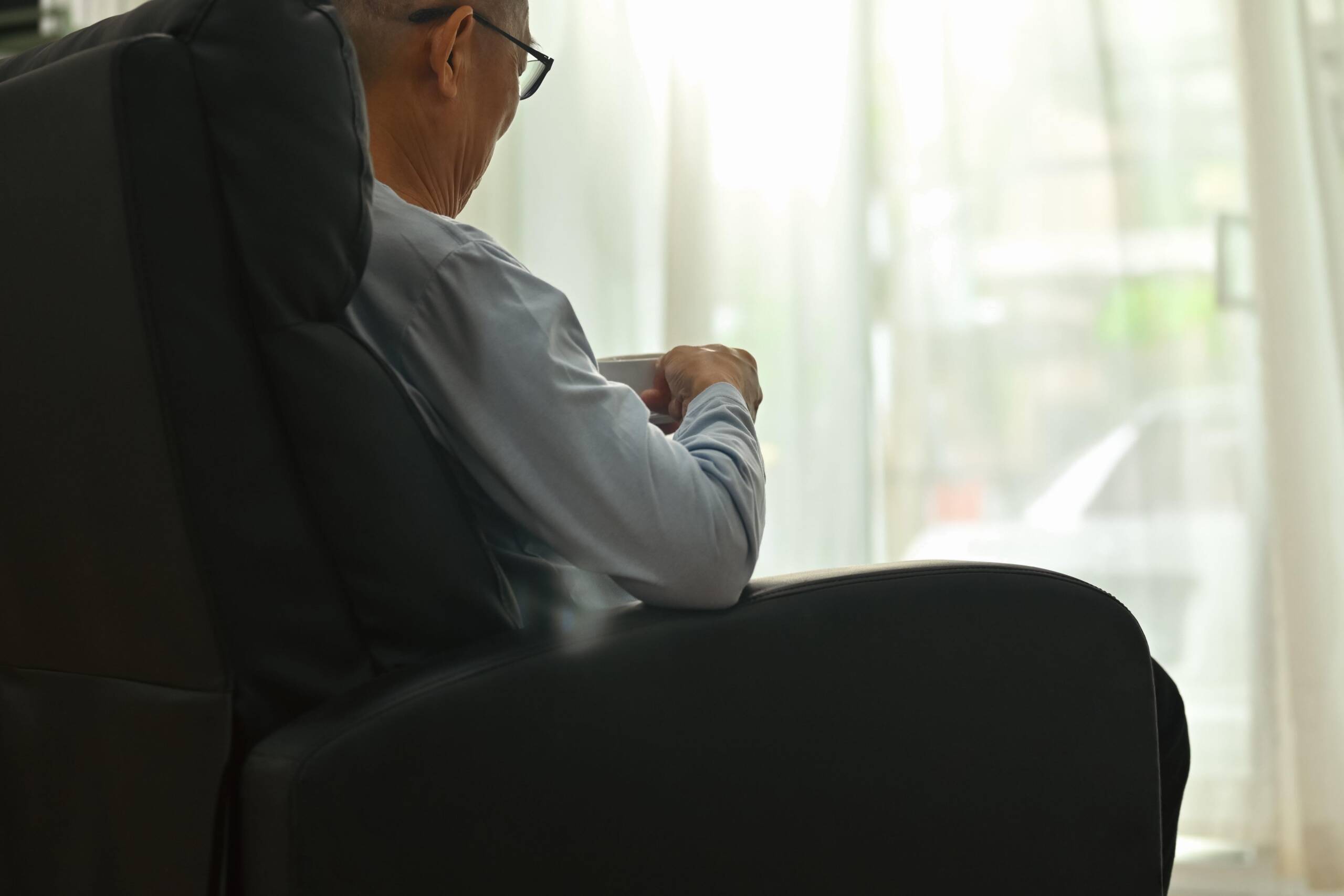 Älterer Mann mit Brille sitzt in einem dunklen Ledersessel und hält eine Tasse in der Hand. Er ist von schräg hinten zu sehen und blickt zu einem hellen Fenster mit weißen Vorhängen. Im Hintergrund steht ein Tisch mit Büchern und einer kleinen Pflanze.